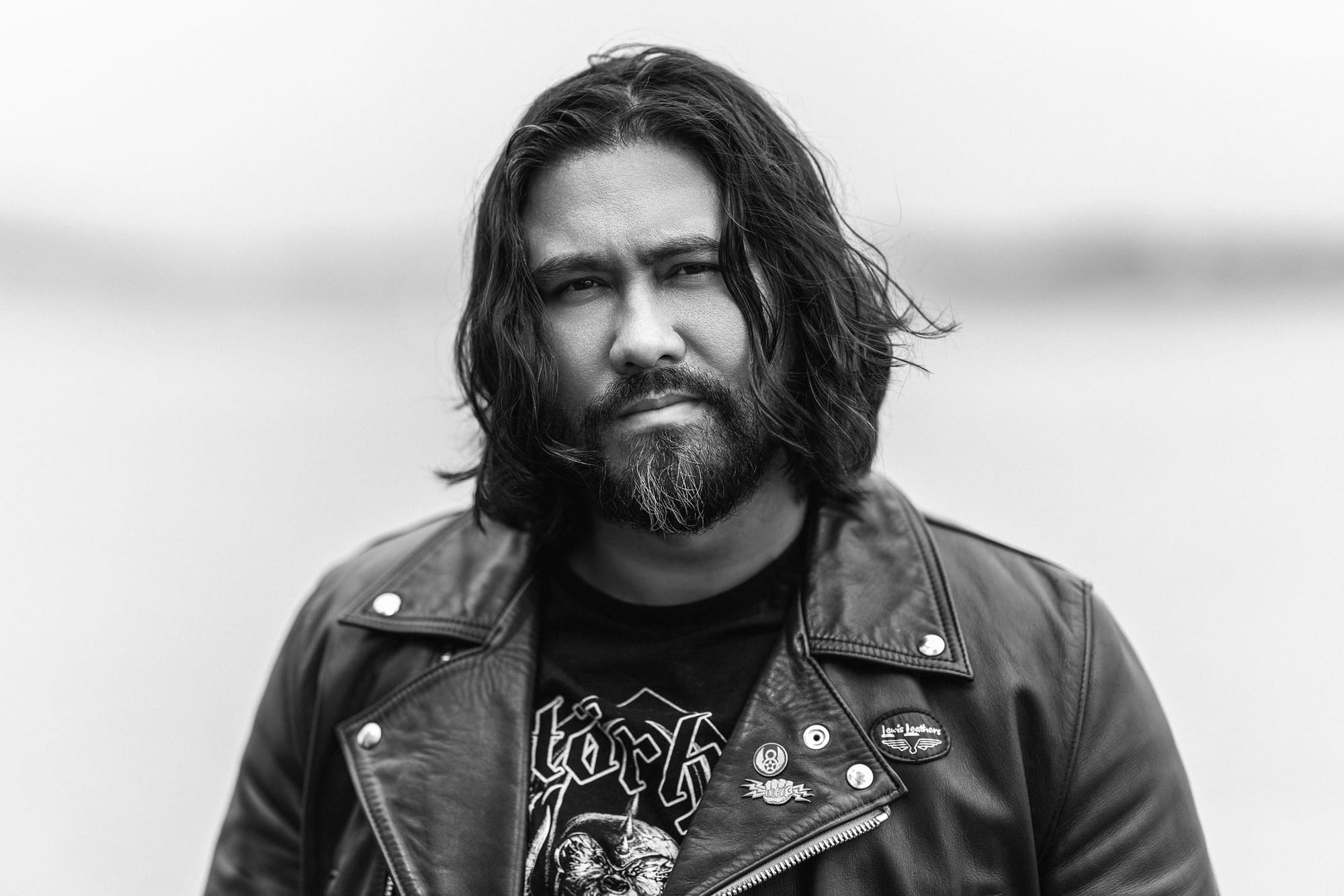 Black and white photograph depicts a mixed race male with long hair and a beard wearing a leather jacket.